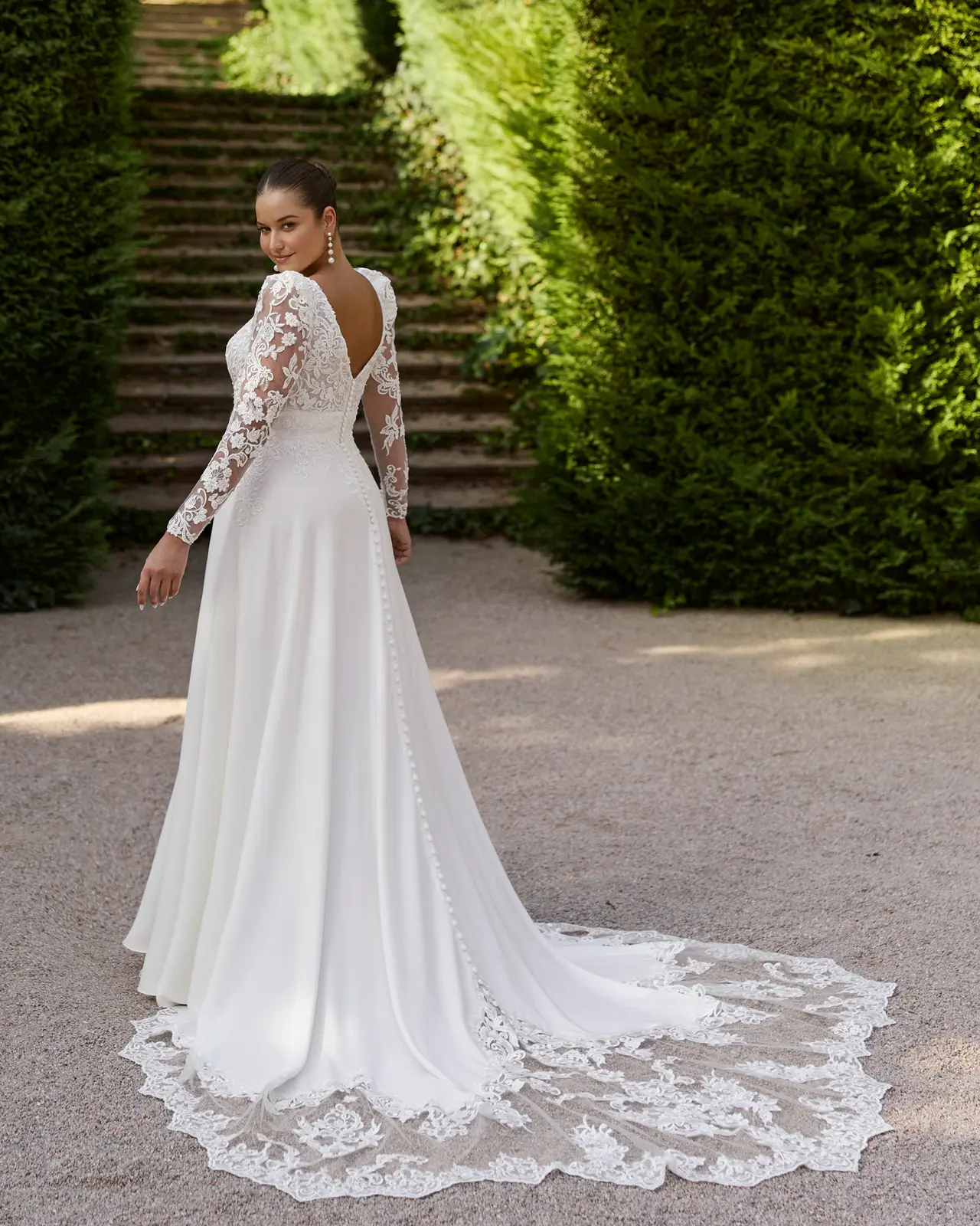 Vista posteriore di un abito da sposa con scollatura a V sulla schiena, maniche decorate e strascico bordato da motivi floreali.